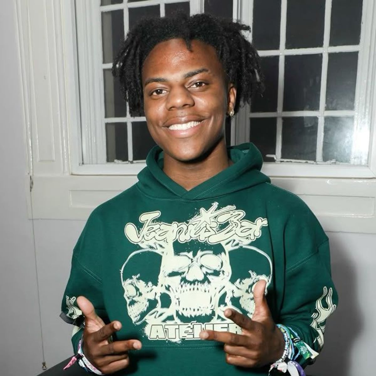 A smiling young person with short dreadlocks wears a green hoodie with skull graphics, standing indoors in front of a white window, making finger-gun gestures with both hands.
