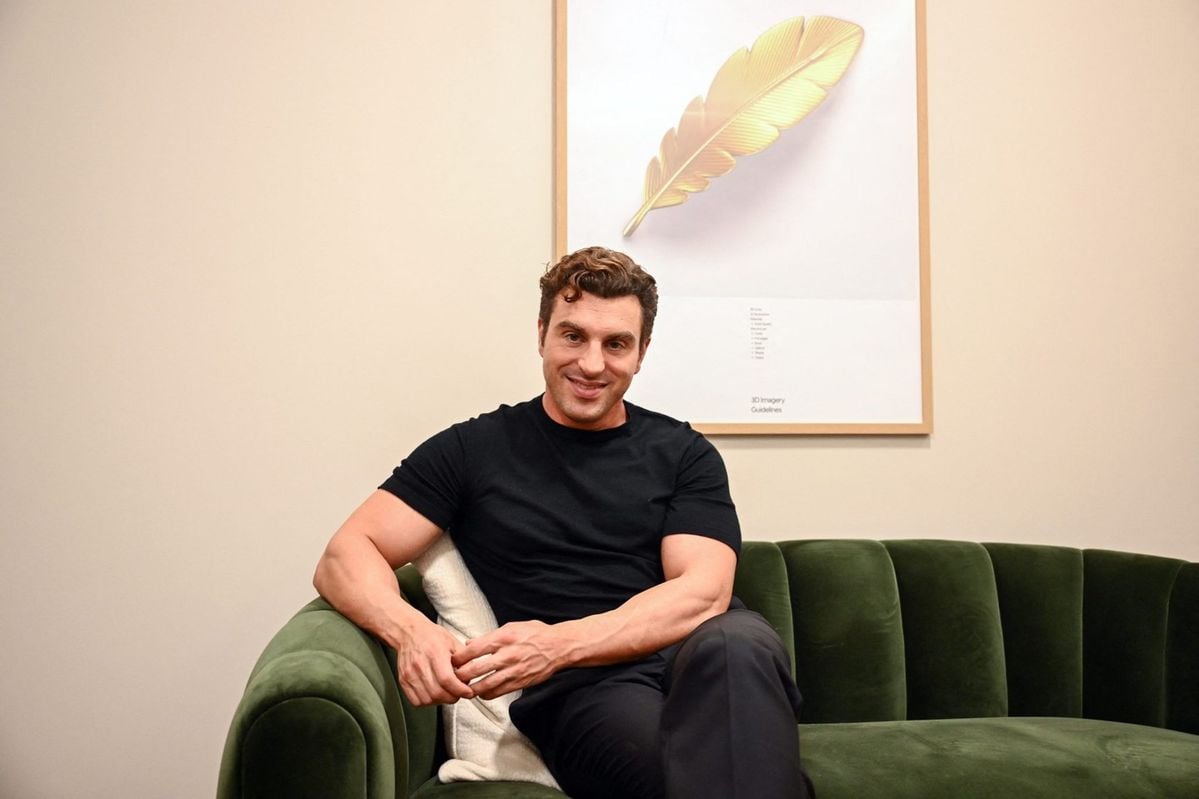 A man with short curly hair sits on a green velvet sofa, smiling, wearing a black t-shirt and dark pants. Behind him is a framed picture of a golden feather on a light-colored wall.