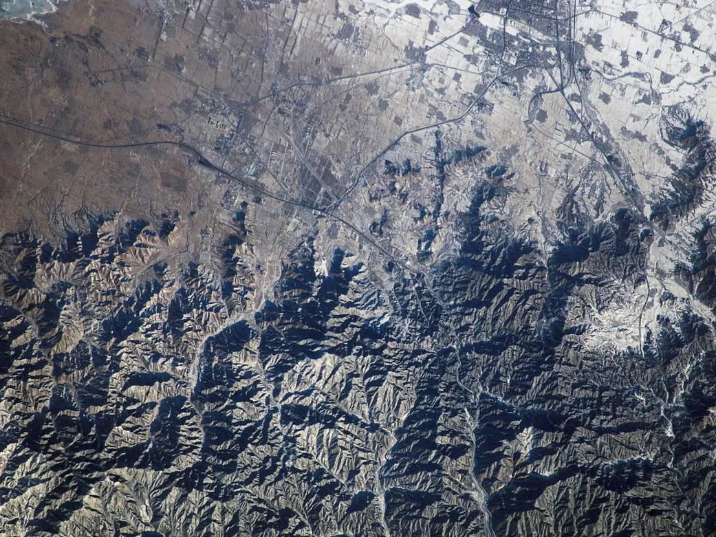 A satellite view showing a rugged, mountainous region transitioning into flat, agricultural land, with a network of roads and small settlements visible. Some mountain peaks and fields are dusted with snow.
