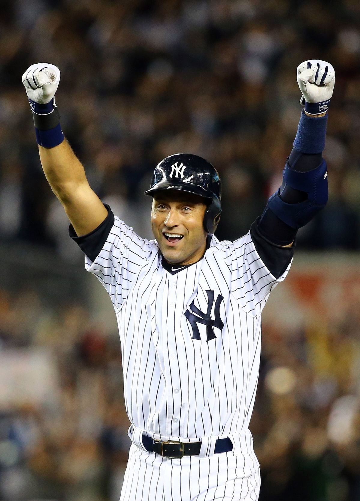 A baseball player in a New York Yankees uniform raises both arms in celebration, smiling, while standing on the field with a crowd in the background.