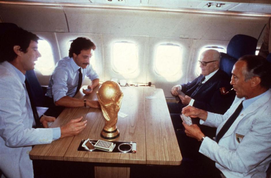 Four men sit around a table on an airplane, playing cards. A FIFA World Cup trophy is placed on the table, along with a wallet and cards. Sunlight shines through the windows in the background.