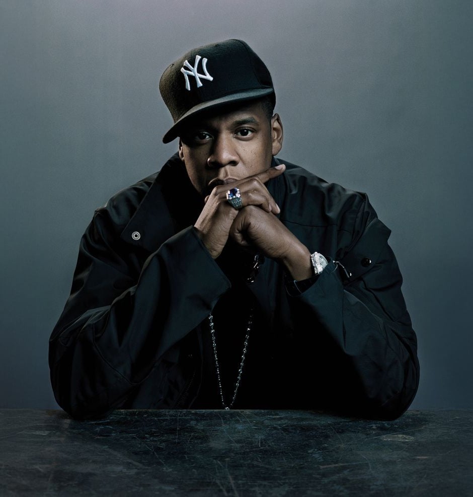 A man wearing a black New York Yankees cap and a dark jacket sits at a table, resting his chin on his hands with fingers interlaced, looking directly at the camera against a plain background.