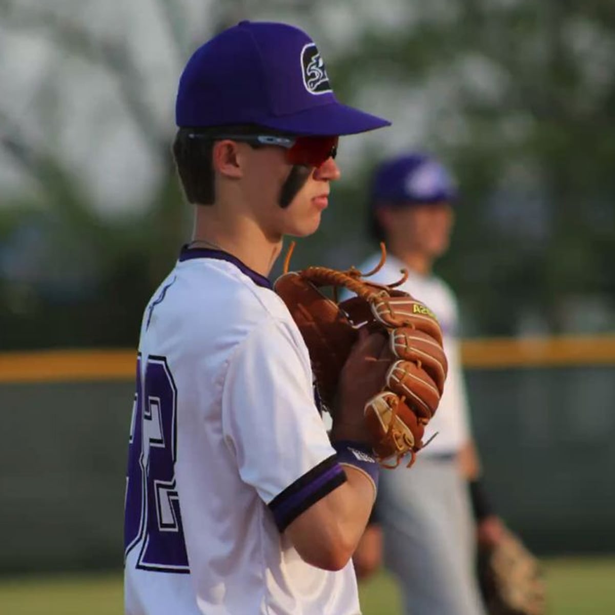 A baseball player in a white and purple uniform and hat stands ready with a glove, wearing eye black and sunglasses, on a field with another player blurred in the background.