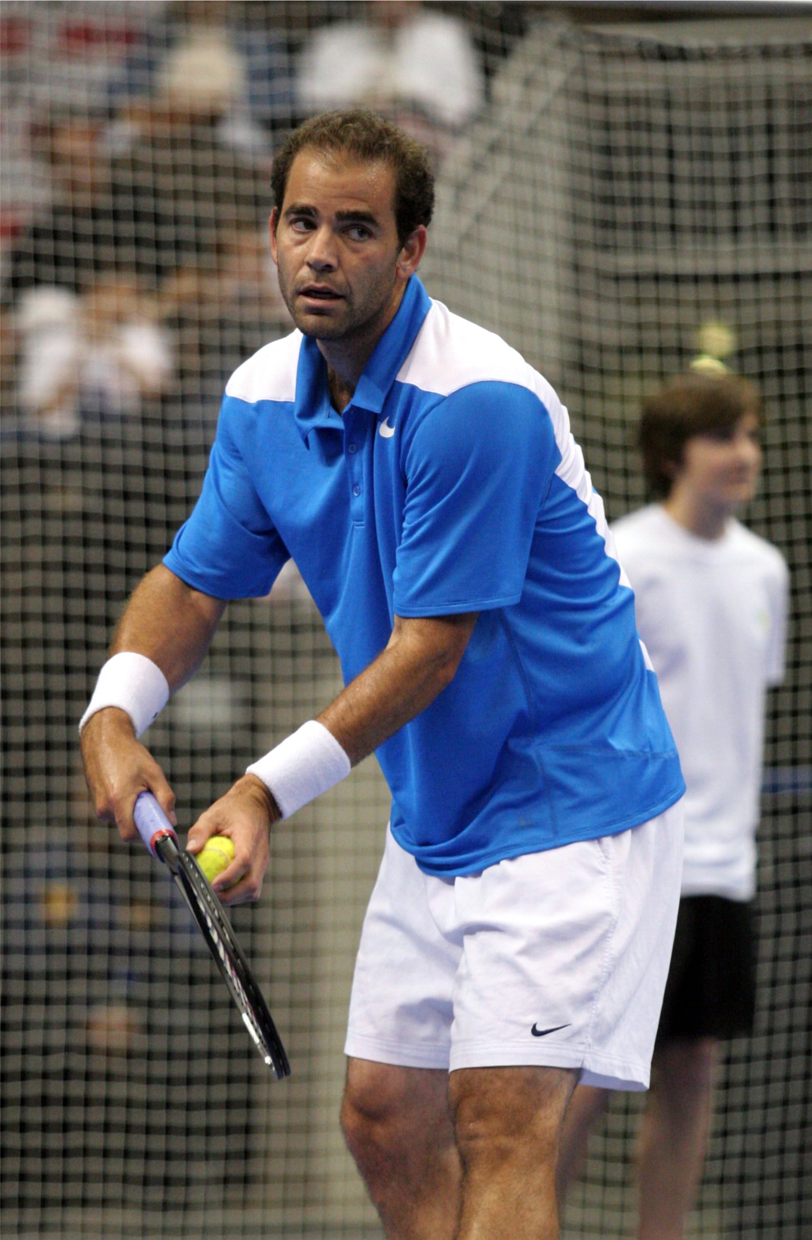A male tennis player in a blue and white outfit prepares to serve on an indoor court, holding a racket in one hand and a tennis ball in the other, with a net and another person blurred in the background.