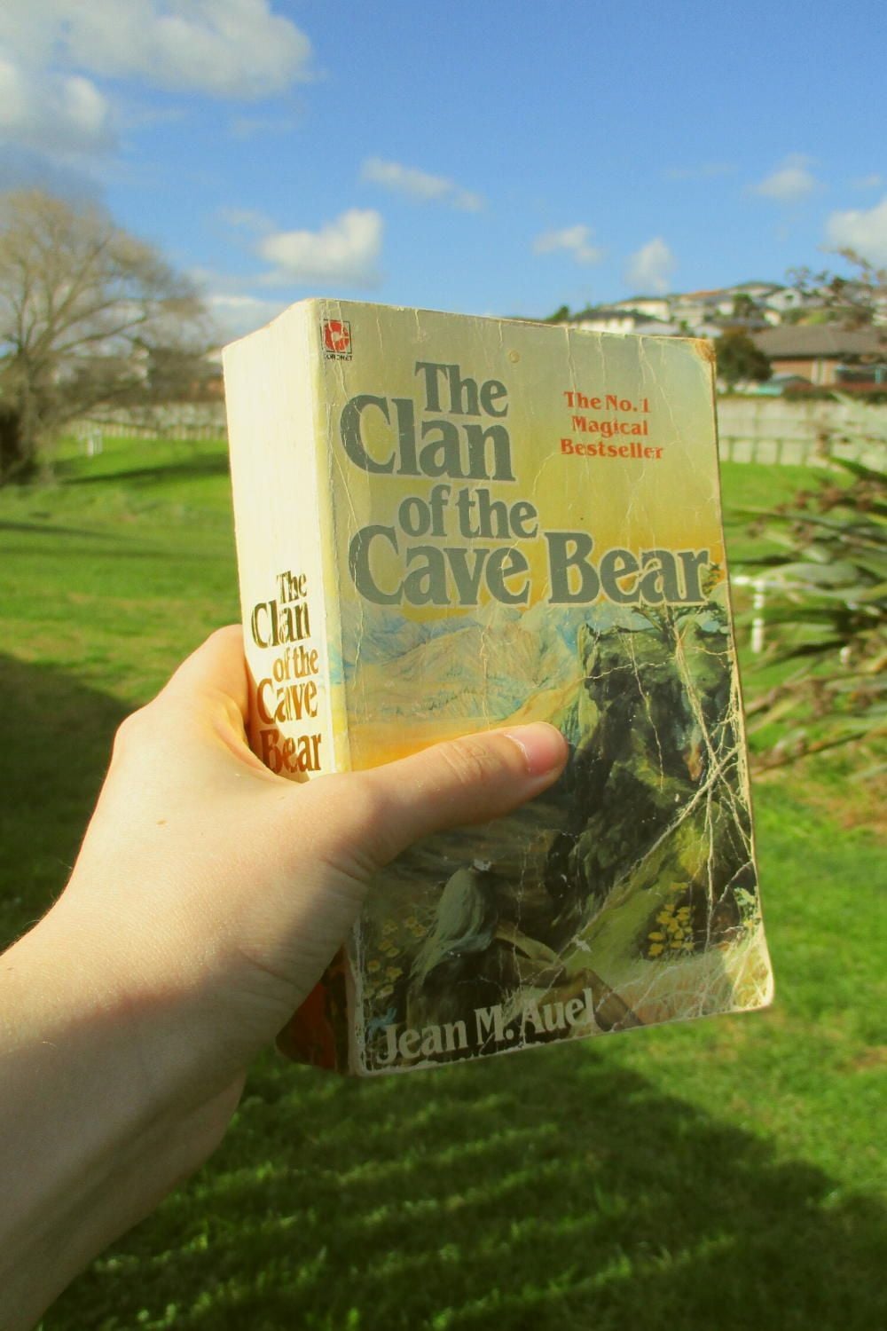 A hand holding a worn paperback copy of “The Clan of the Cave Bear” by Jean M. Auel outdoors, with green grass, trees, and a partly cloudy blue sky in the background.