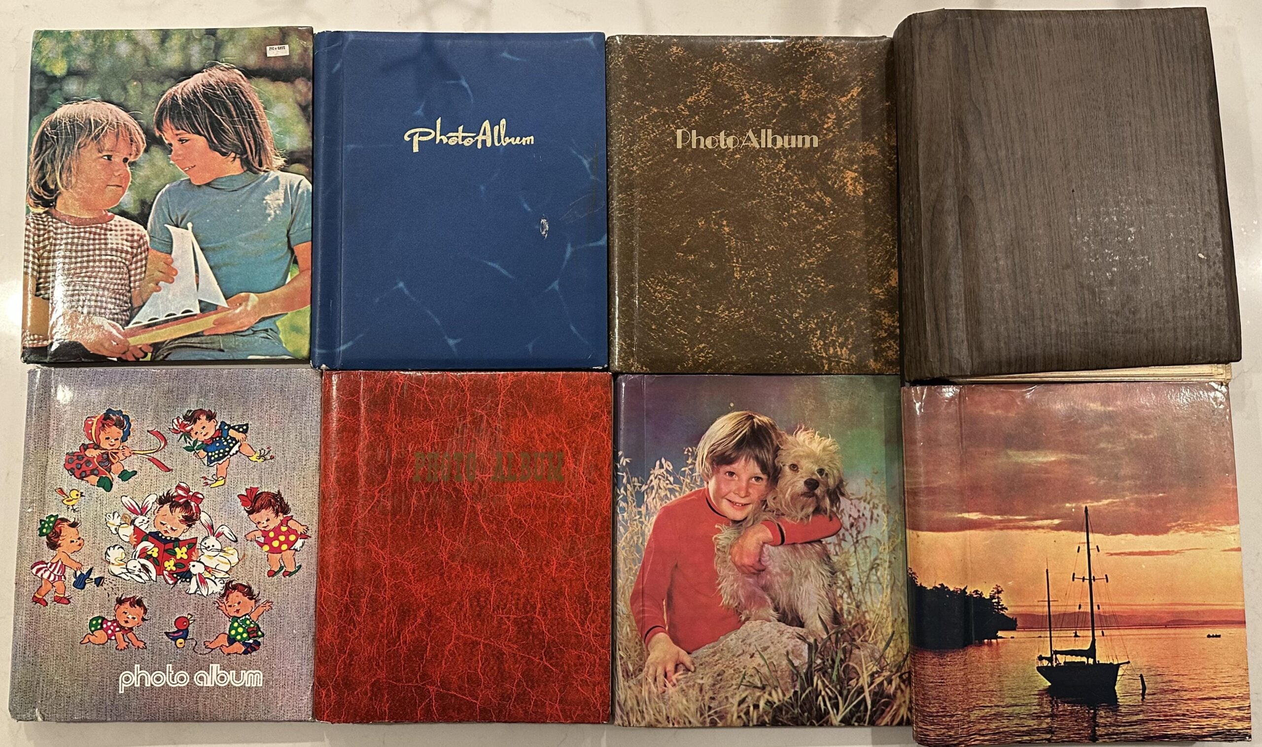 Eight vintage photo albums are arranged in two rows. Covers feature children, colorful cartoon characters, a boy with a dog, a sailboat at sunset, and various colors and designs, including blue, red, and brown.