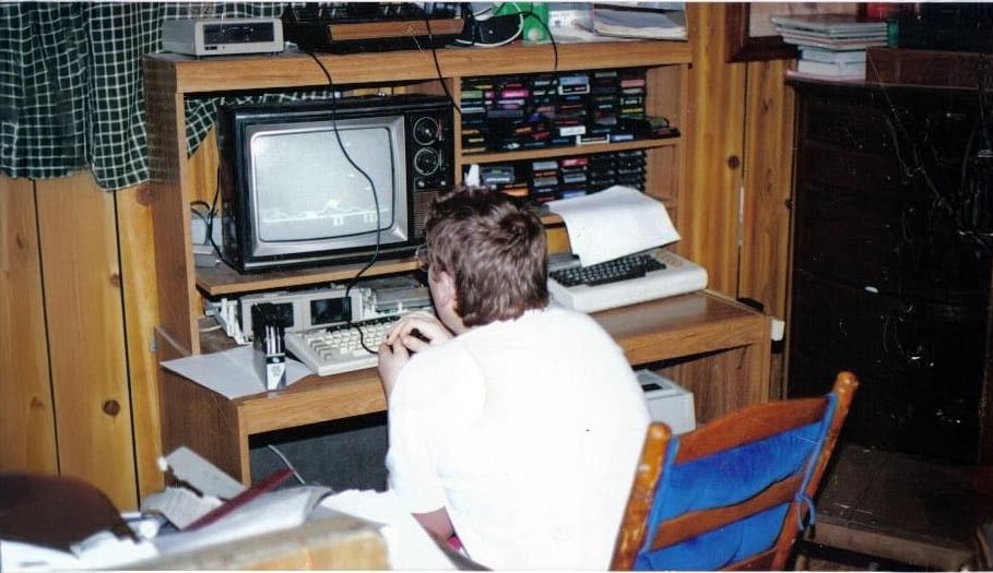 A person sits at a wooden desk using a vintage computer and CRT monitor, surrounded by floppy disks and papers, in a wood-paneled room with shelves and electronic equipment.