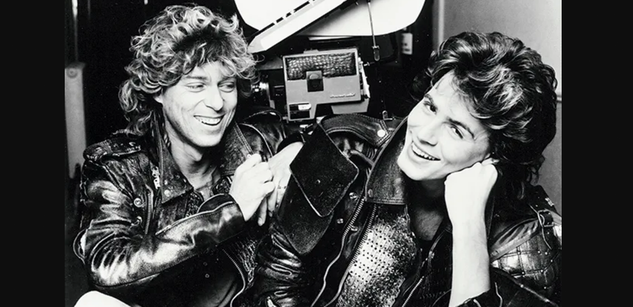 Two young men with curly hair, wearing shiny leather jackets, smile and laugh together in front of film equipment in this black and white photo.