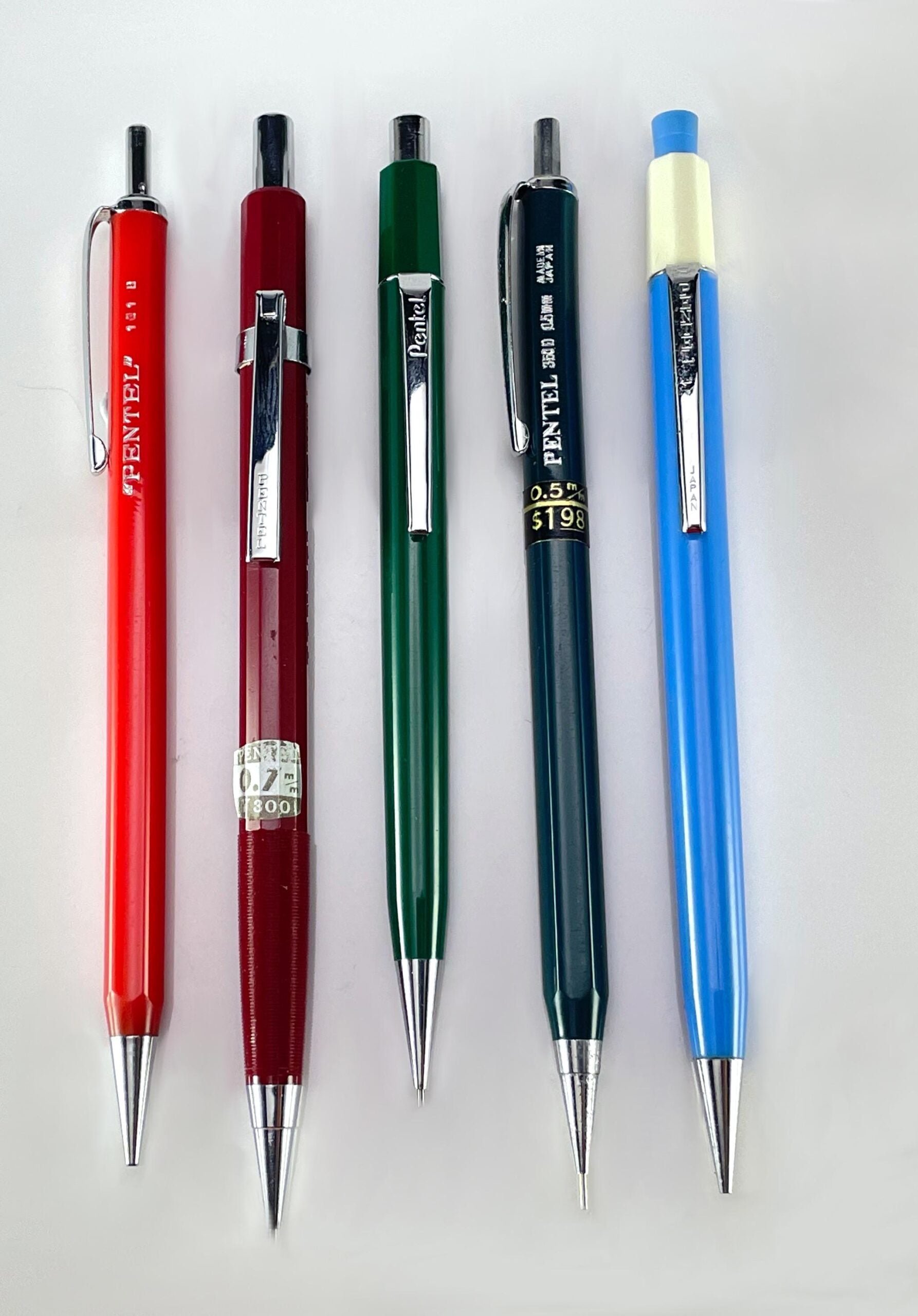 Five mechanical pencils in red, burgundy, green, dark green, and blue with silver clips and tips are lined up vertically on a white background. The blue pencil has an eraser visible at the top.