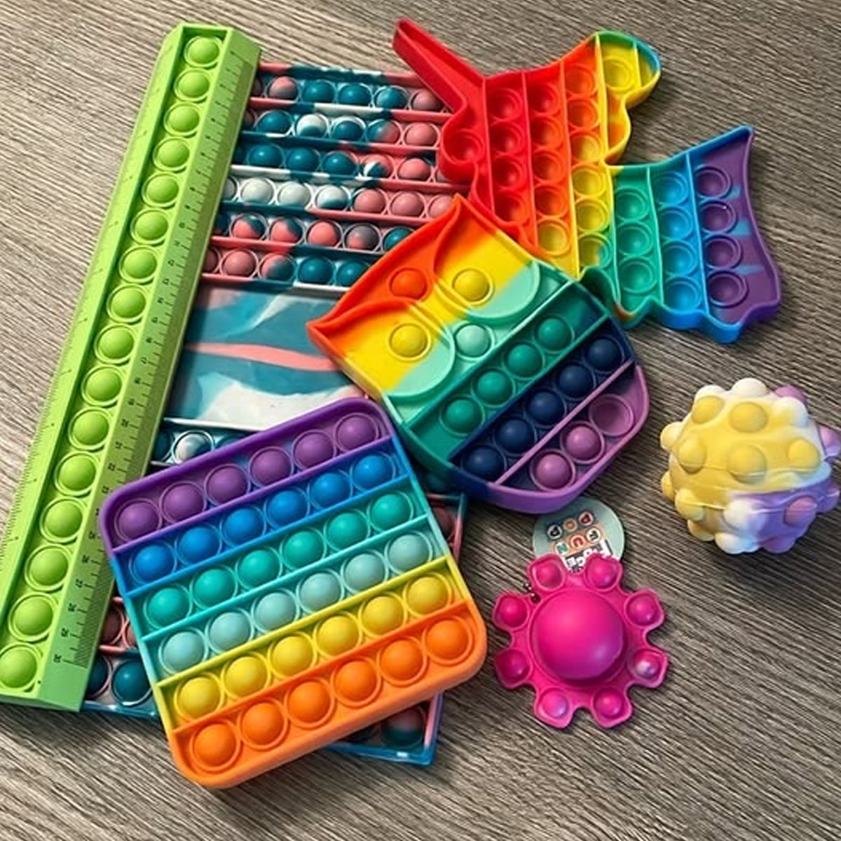 A collection of colorful pop-it fidget toys in various shapes, including a square, owl, among others, plus a ruler, a textured ball, and a purple and pink spinner, all on a wooden surface.