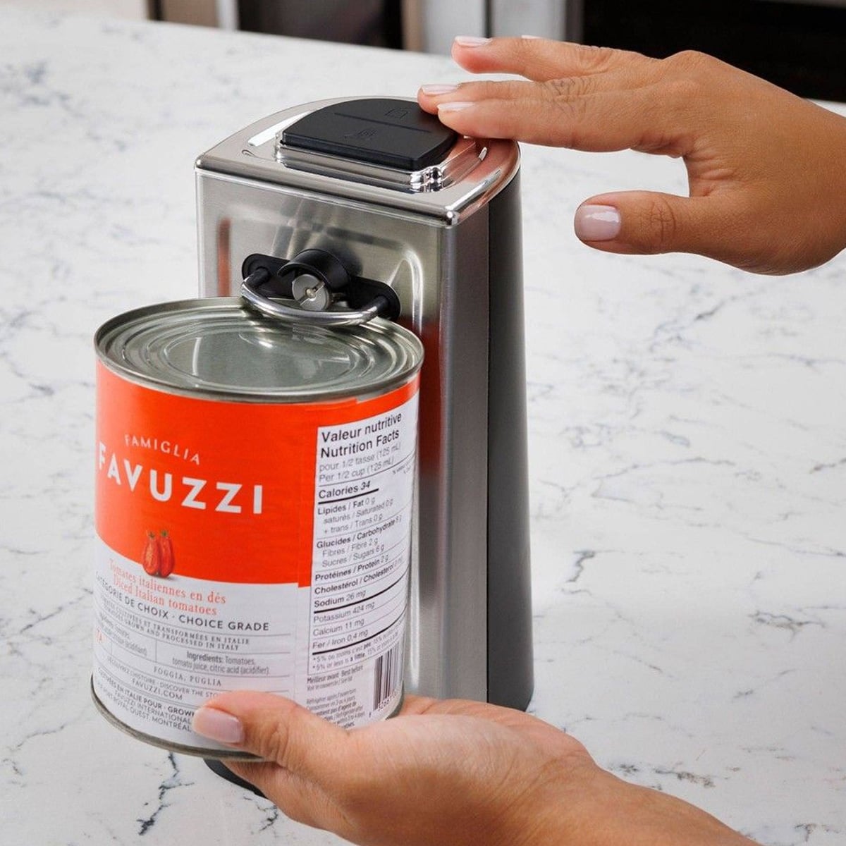 A person uses an electric can opener on a kitchen counter to open a large can of Favuzzi tomatoes, holding the can in place while pressing the opener’s button.