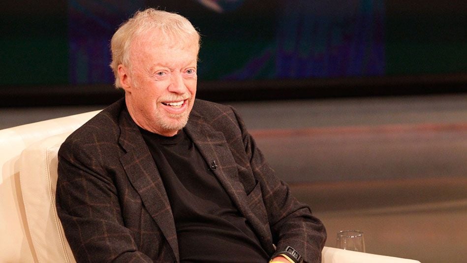 An older man with light hair and a short beard, wearing a dark plaid blazer and black shirt, sits smiling on a light-colored chair in a studio setting.