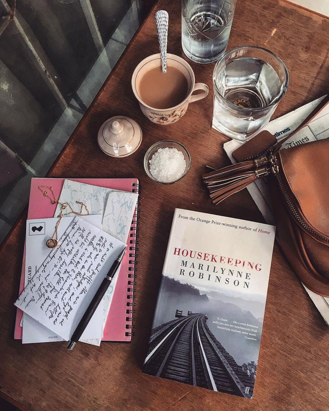 A wooden table with a cup of coffee, a glass of water, a brown purse, a book titled "Housekeeping" by Marilynne Robinson, a handwritten letter, a notebook, a pen, and a small bowl of sugar.