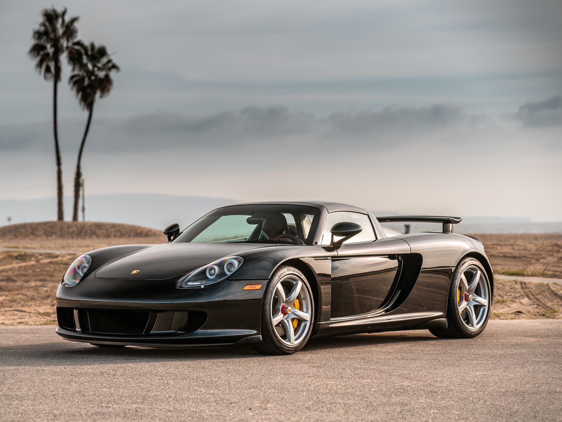 A black Porsche Carrera GT sports car parked on a road near a beach, with palm trees and a cloudy sky in the background.