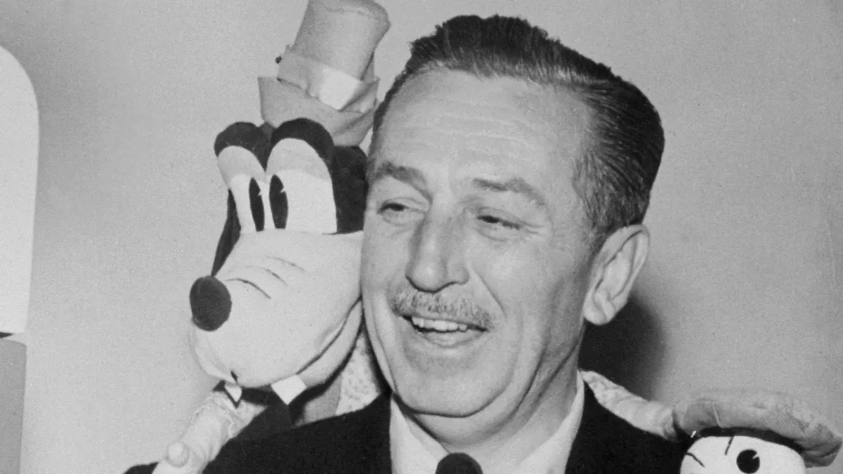 A smiling man with a mustache stands in front of a large cartoon character statue wearing a top hat, likely at an event or celebration. The photo is in black and white.