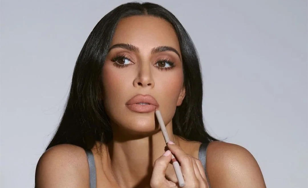 A woman with long dark hair applies nude lip liner to her lips while looking slightly to the side. She wears a gray top and has soft, natural makeup with defined brows and a matte finish.