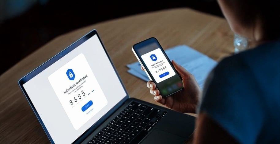 A person holds a smartphone displaying a verification code while sitting at a desk with a laptop that shows the same code for account authentication.