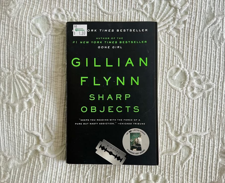 A paperback copy of "Sharp Objects" by Gillian Flynn rests on a white textured fabric. The black cover features neon green text and an image of a razor blade at the bottom. Price stickers are on the top left corner.