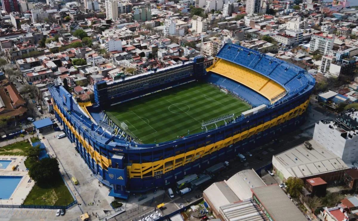 Aerial view of a large, blue and yellow football stadium surrounded by urban buildings and streets, with green grass on the field and empty stands.