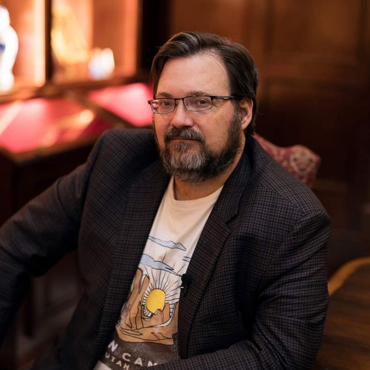 A bearded man with glasses sits in a warmly lit room, wearing a checked blazer over a printed T-shirt. He looks at the camera with a relaxed expression. The wooden background adds a cozy atmosphere.
