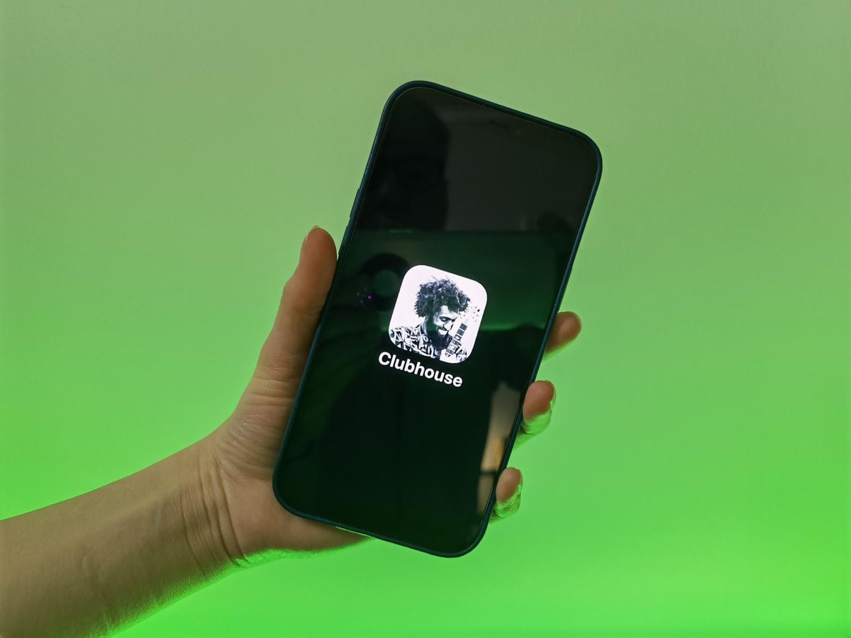 A hand holds a smartphone displaying the Clubhouse app logo on the screen, with a green gradient background behind it.