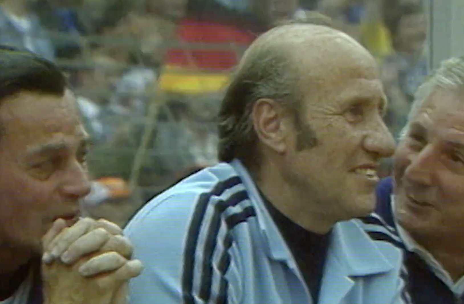 Three older men sit closely together, smiling and talking. The central man wears a light blue jacket with dark stripes. The background is blurred, showing a crowd and a flag with red and yellow stripes.