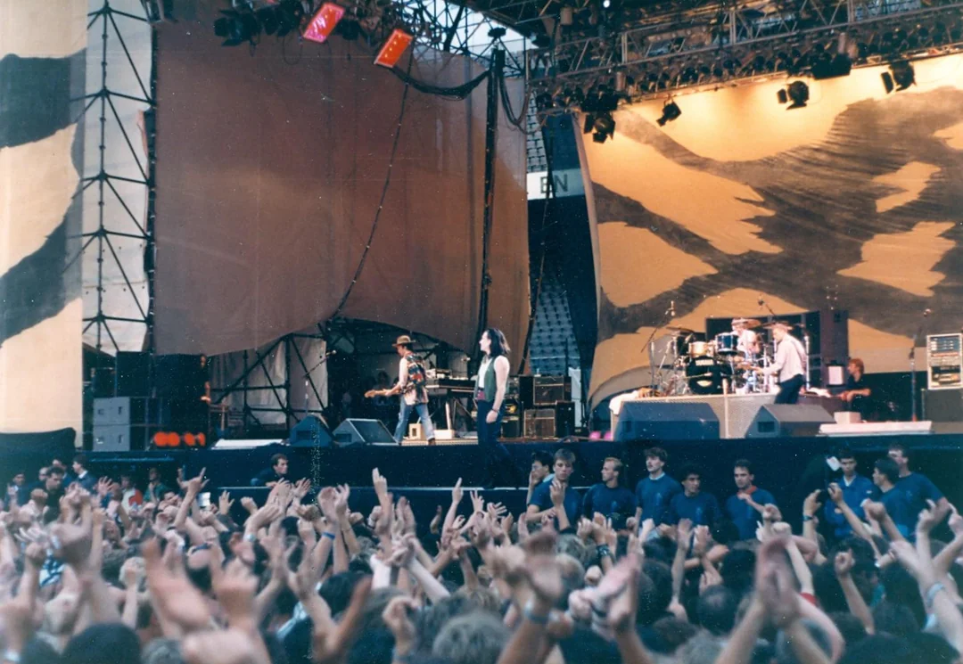 A band performs on an outdoor stage with dramatic lighting and abstract backdrops, while a large crowd of fans raises their hands and cheers in front of the stage.
