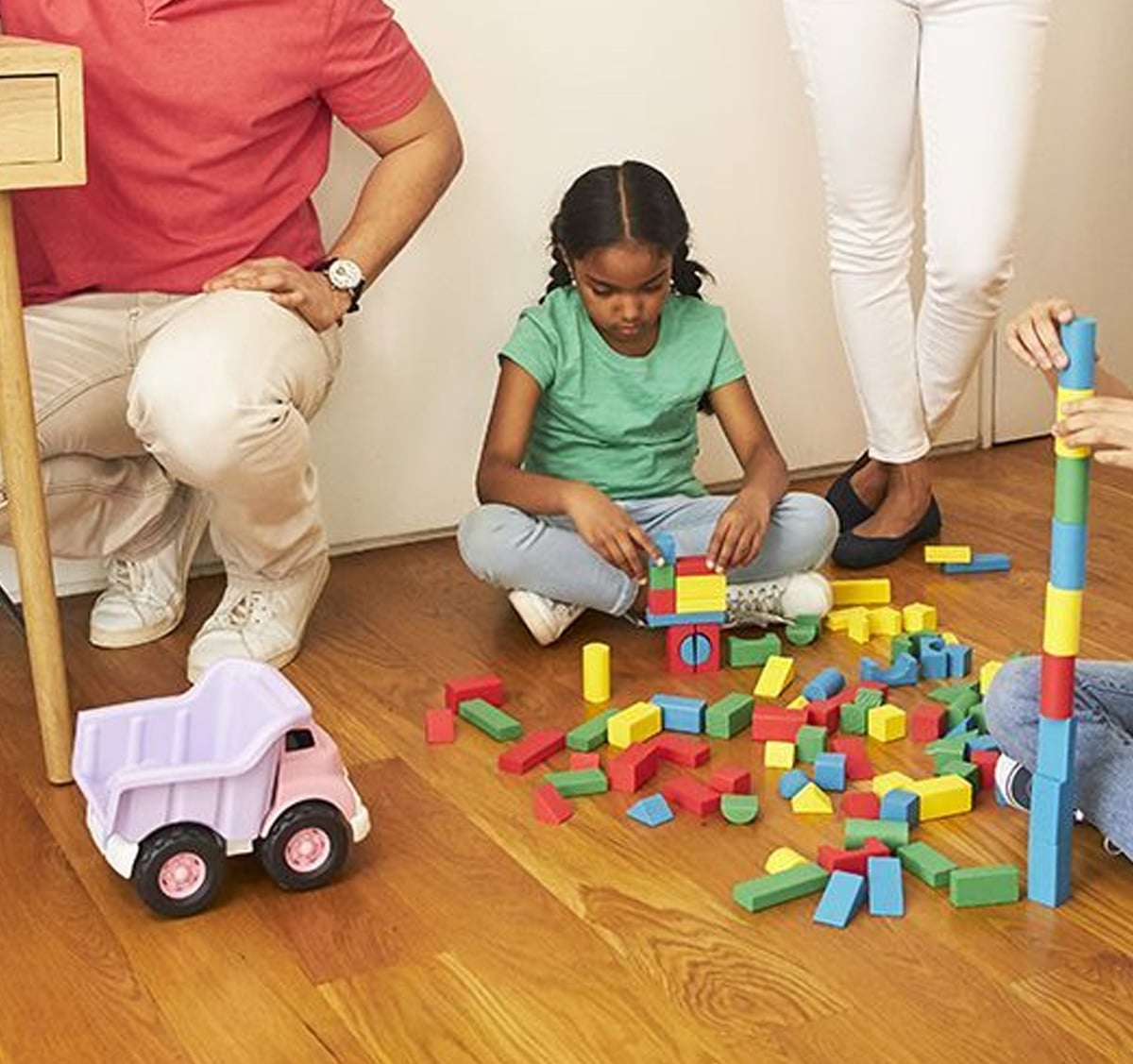 A child sits on a wooden floor surrounded by colorful building blocks, assembling a structure. Nearby, adults stand and kneel, and a purple toy truck is visible on the floor.