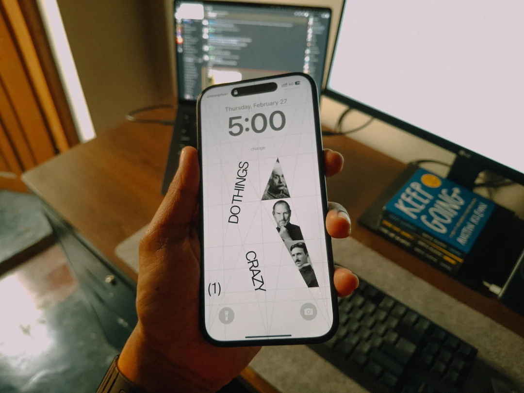 A hand holds a smartphone displaying a lock screen with the text "DO THINGS CRAZY" and black-and-white portraits. In the background are a computer monitor, keyboard, and two stacked books on a desk.
