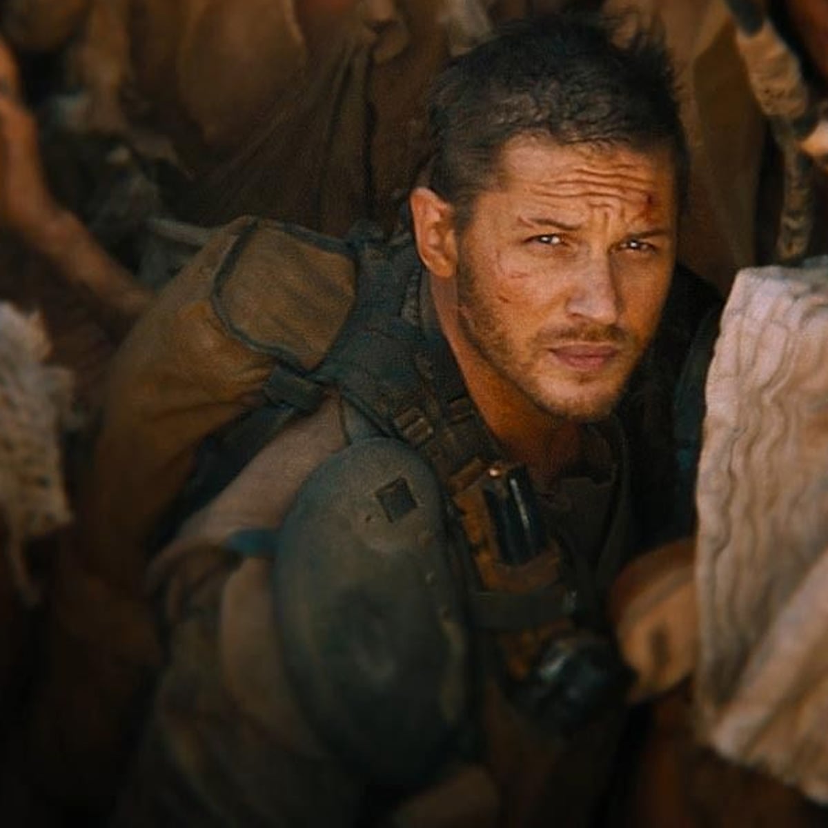 A man with short hair and a rugged, dirty face wears worn-out clothing and gear, crouching among a crowd in a tense, desert-like environment. Harsh lighting highlights his serious, intense expression.