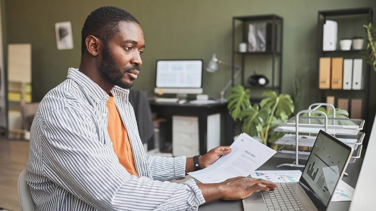 A man sits at a desk in an office, holding documents in one hand and using a laptop with the other. There are charts on the laptop screen and paperwork on the desk. Shelves and plants can be seen in the background.