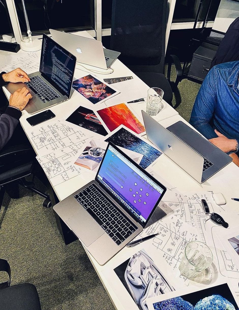 A cluttered table with three laptops, scattered photos, sketches, documents, pens, and glasses of water. Two people are partially visible, one in a blue shirt, both working at their laptops.