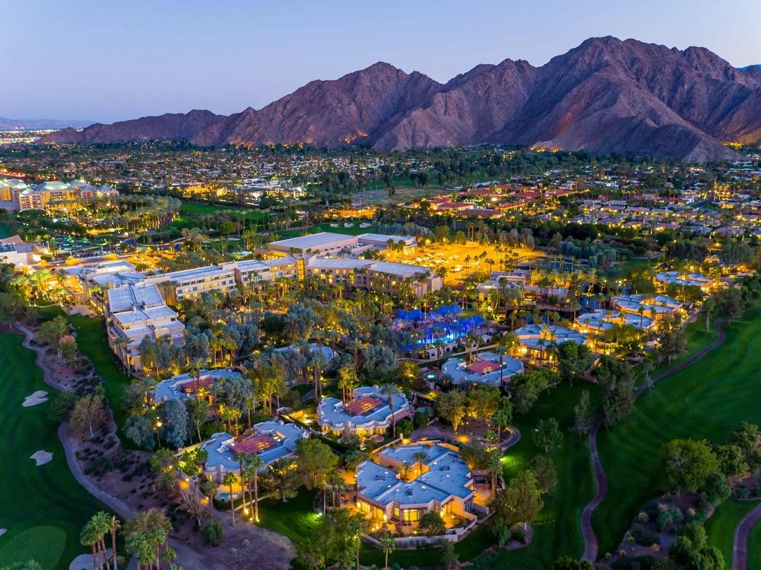 Aerial view of a resort with illuminated buildings, pools, and palm trees at dusk, set against a backdrop of tall mountains and a sprawling city in the distance.