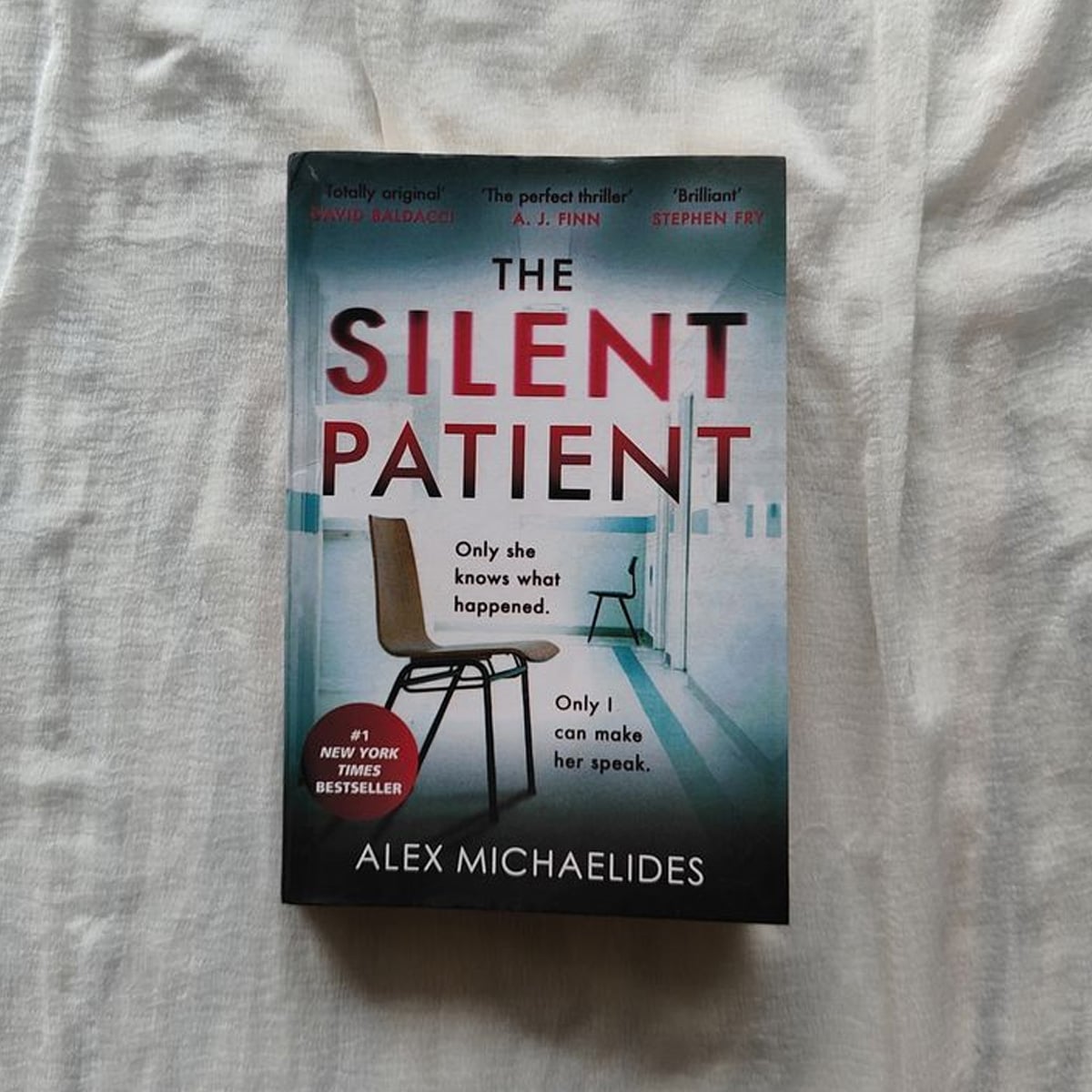 The book "The Silent Patient" by Alex Michaelides is placed on a white fabric background. The cover features a solitary chair facing a window, with bold red and white text and a New York Times Bestseller sticker.
