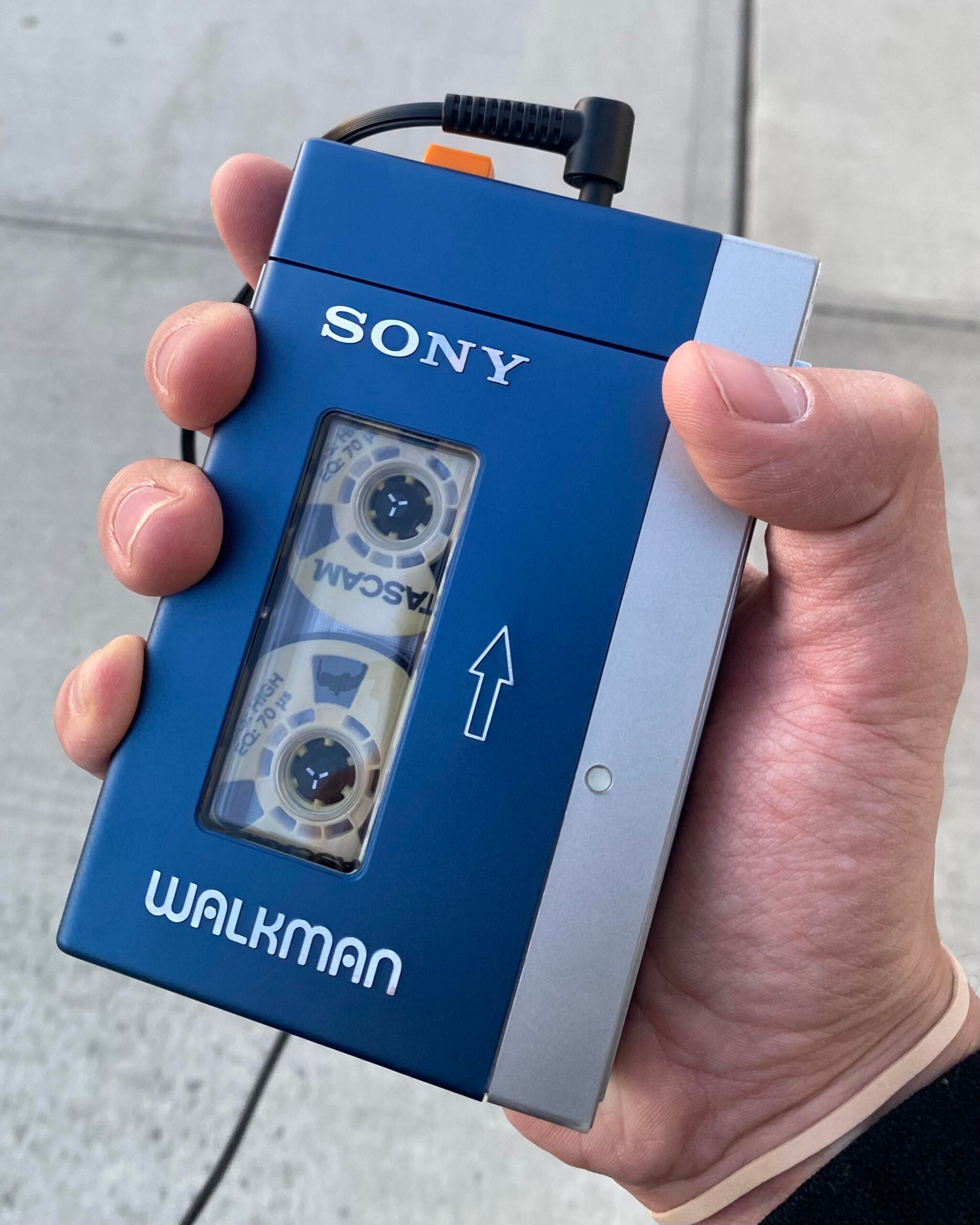 A hand holds a blue and silver Sony Walkman cassette player with a visible cassette tape inside, shown outdoors on a concrete sidewalk.