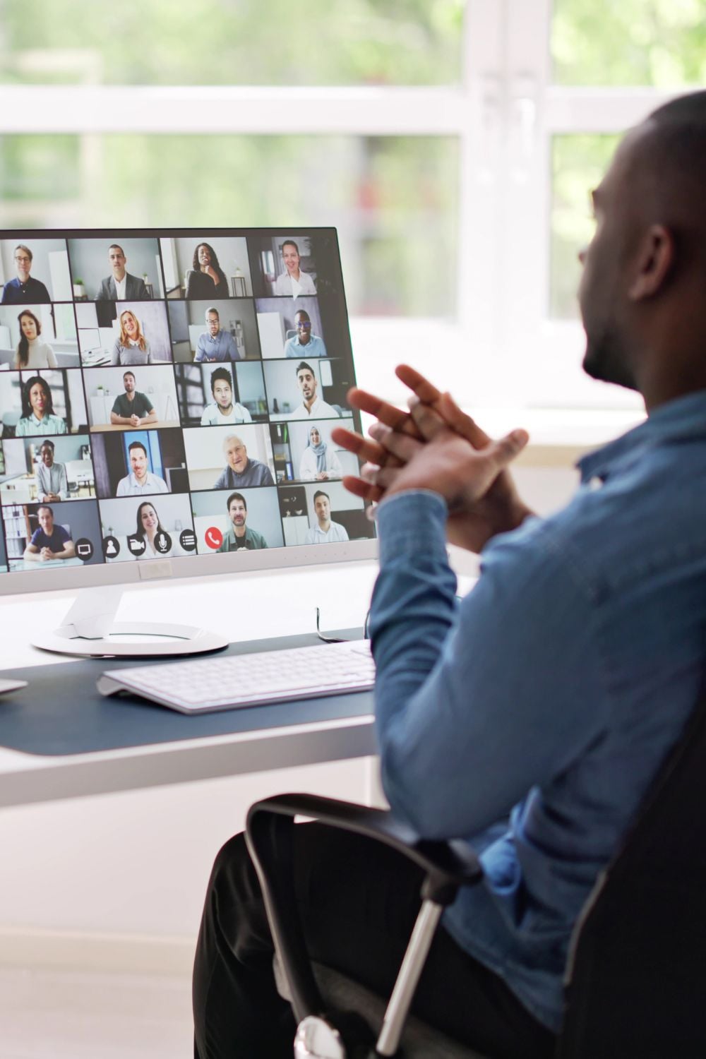 A person sits at a desk and participates in a video conference call on a computer monitor, displaying a grid of many people in small windows. The setting appears to be a bright home office.