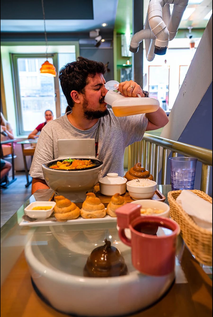 A man sits at a table in a restaurant, drinking from a large bottle of juice. In front of him are several dishes styled to look like poop, including brown swirls and mugs shaped like toilets.