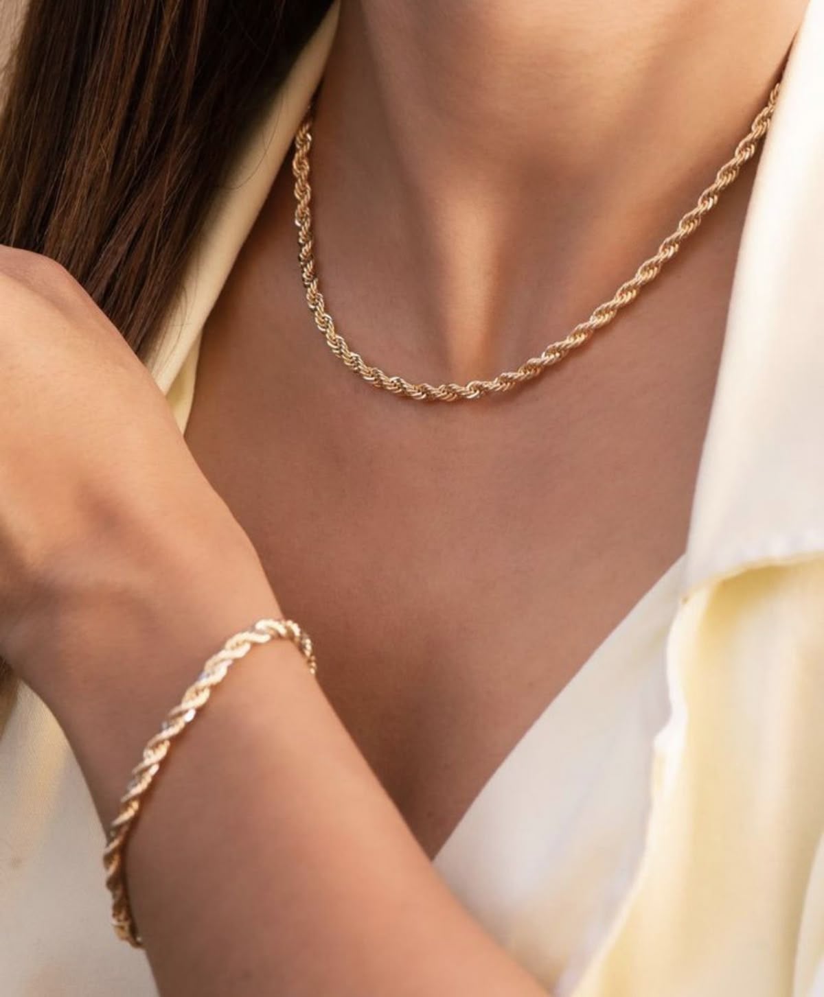 A woman wearing a twisted gold chain necklace and matching bracelet, dressed in a cream-colored blouse. Her hand gently touches her shoulder, highlighting the elegant jewelry set.