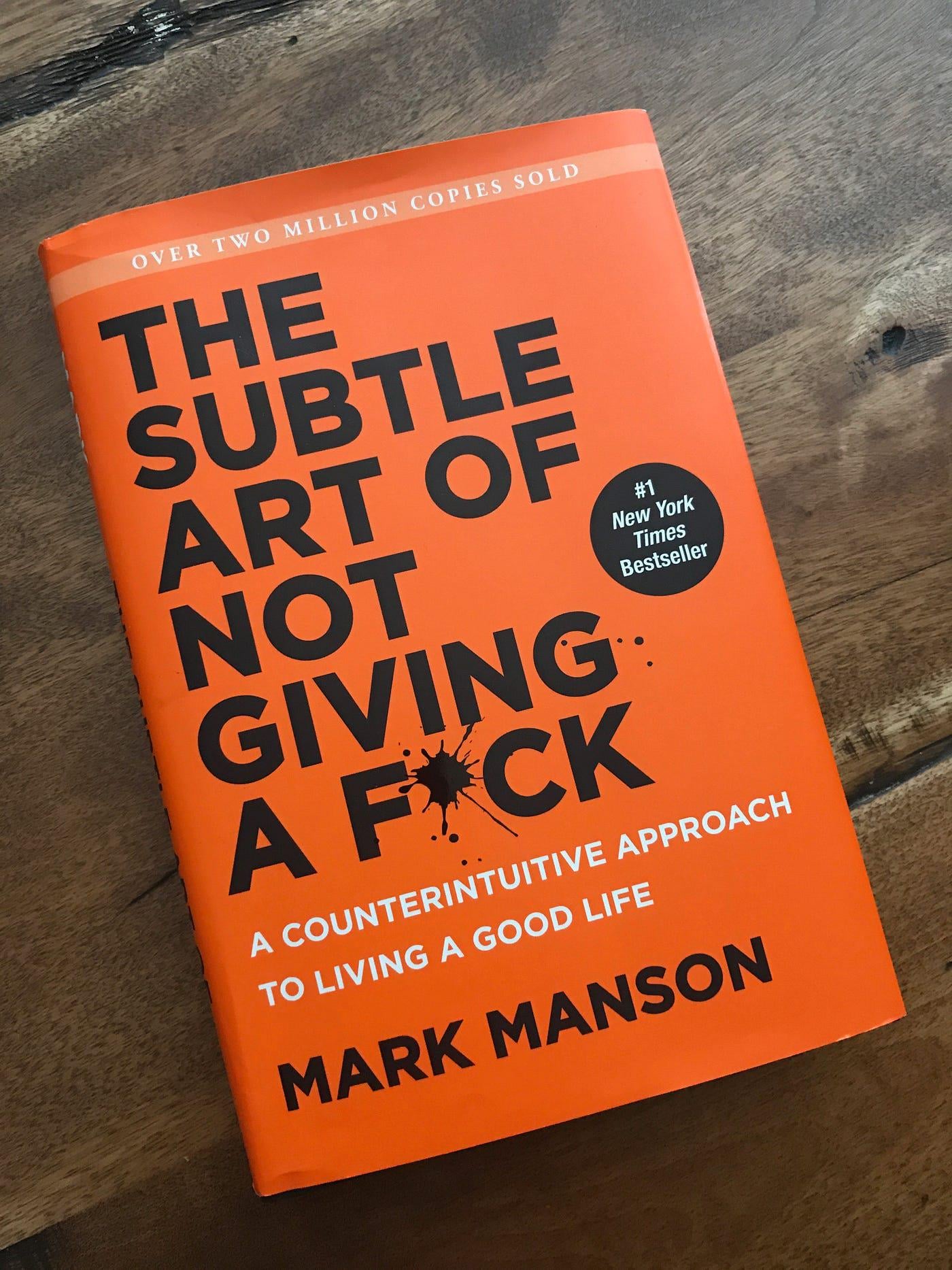 An orange book titled "The Subtle Art of Not Giving a F*ck: A Counterintuitive Approach to Living a Good Life" by Mark Manson rests on a wooden surface. A black sticker notes it's a New York Times bestseller.
