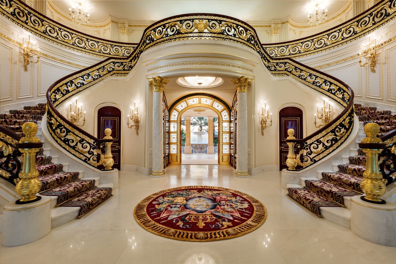A grand staircase with ornate gold railings curves upward on both sides of a luxurious foyer with marble floors, elaborate wall moldings, and a decorative round rug at the center. A chandelier hangs above double doors.
