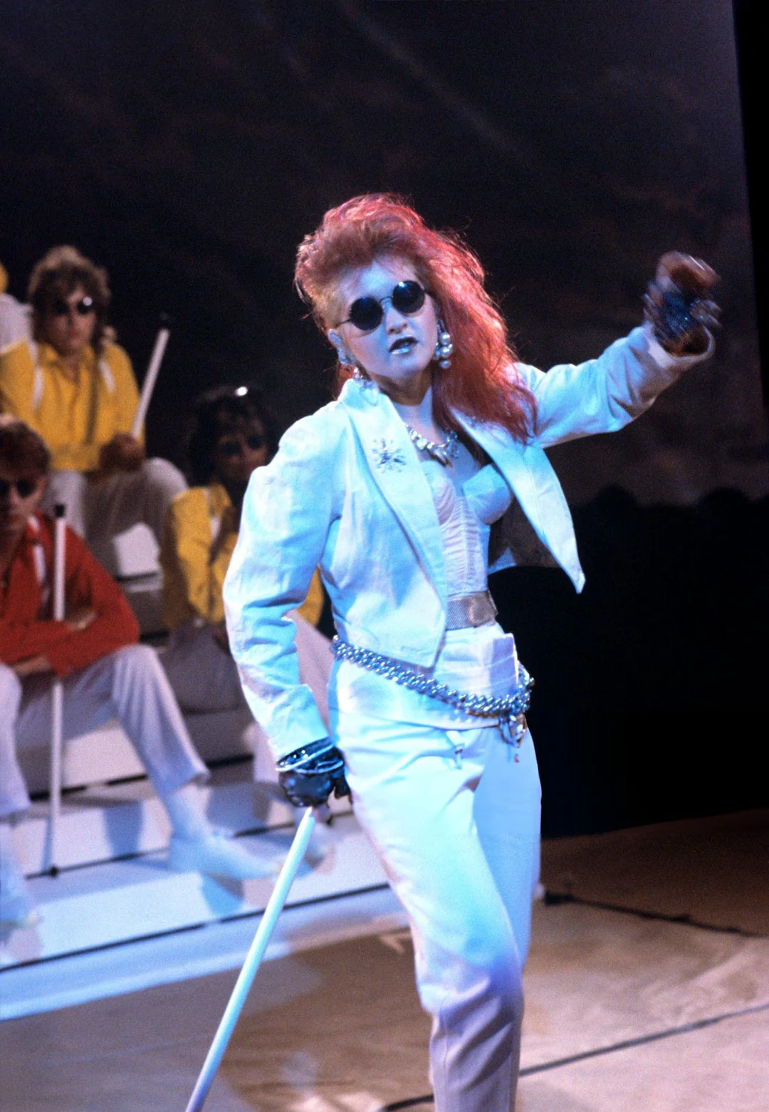 A performer with bright red hair, wearing sunglasses, a white suit, and gloves, sings energetically on stage. People in colorful outfits sit in the background, watching the performance.