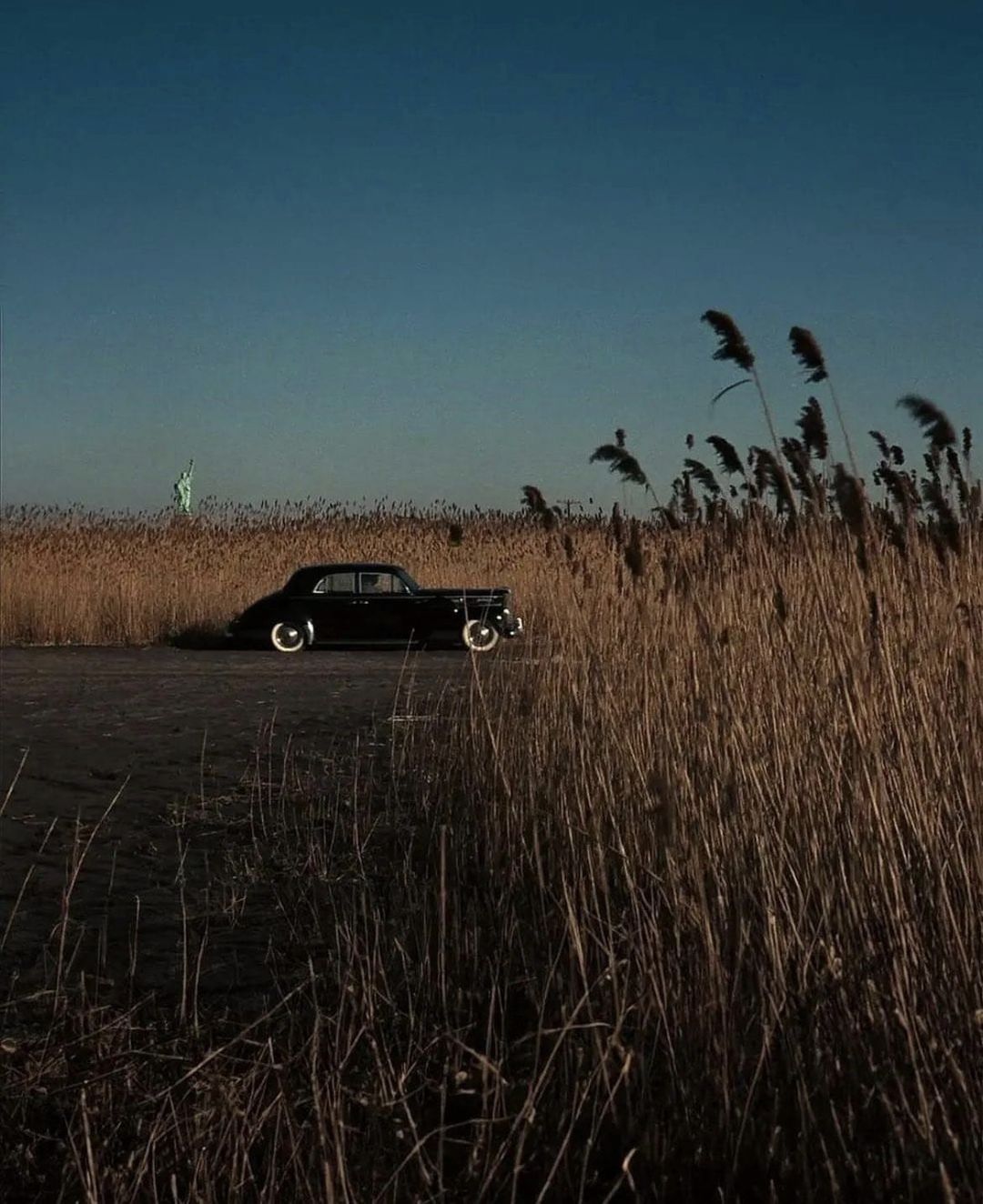 A vintage black car is parked on a dirt road beside tall, golden dry grass under a deep blue sky, with the Statue of Liberty visible in the distant background.