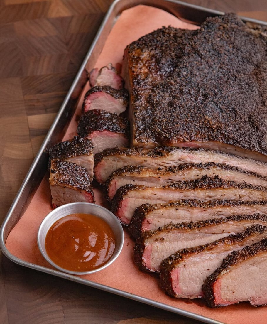 Sliced smoked brisket with a dark crust arranged on a tray lined with parchment paper, served with a small metal cup of barbecue sauce on the side.
