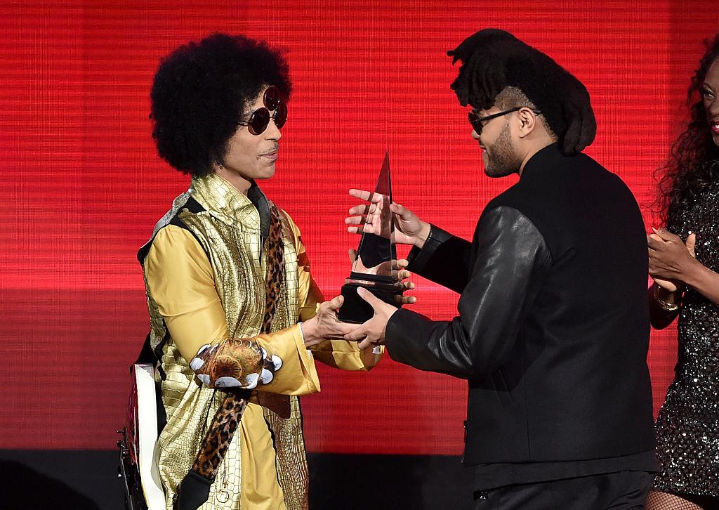 A man in a gold and black outfit with sunglasses hands an award trophy to another man in dark clothing and sunglasses on stage, with a woman clapping in the background.