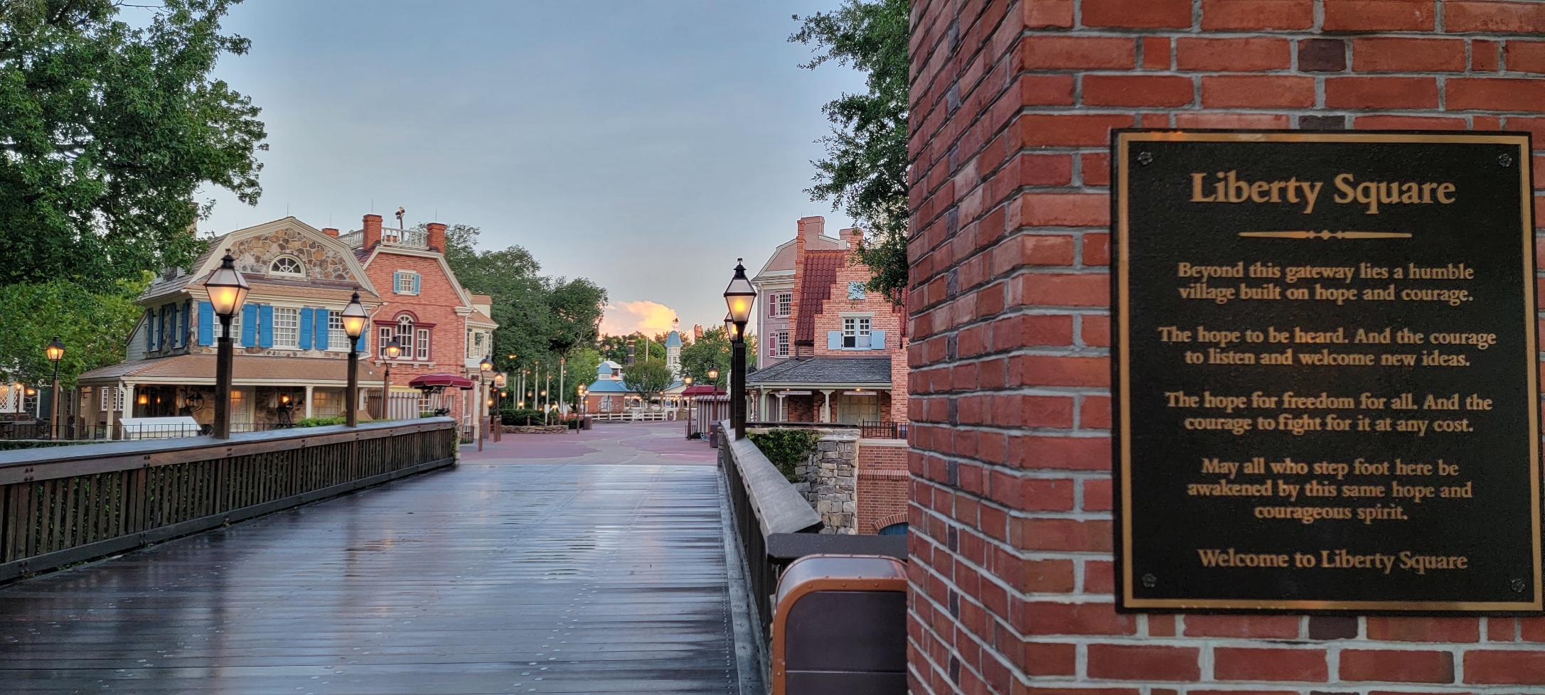 A brick pathway leads into Liberty Square with colonial-style buildings and trees on either side. A plaque on a brick wall displays an inspirational message about hope, courage, and freedom.