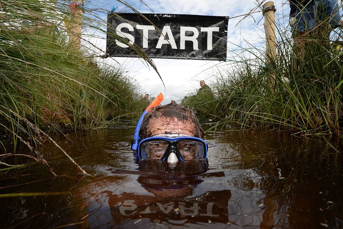 A person wearing a snorkel and goggles is submerged in muddy water at the starting line of an outdoor race, with a "START" sign overhead and grassy banks on either side.