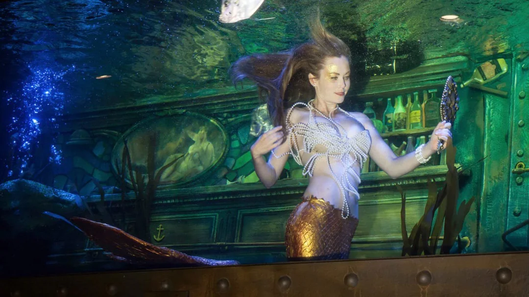 A woman dressed as a mermaid with a pearl top and golden tail poses underwater in front of a bar setting, holding a hand mirror, with her hair flowing around her.