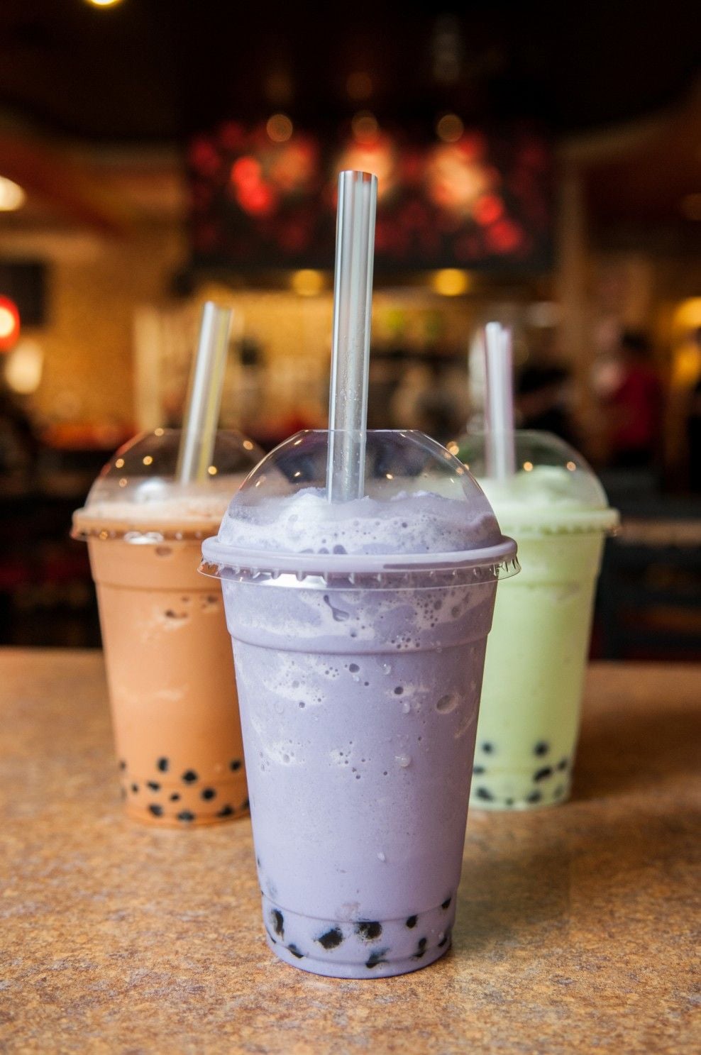 Three bubble tea drinks in plastic cups with dome lids and straws sit on a table. The drinks are purple, green, and brown, each with black tapioca pearls at the bottom. The background is softly blurred.