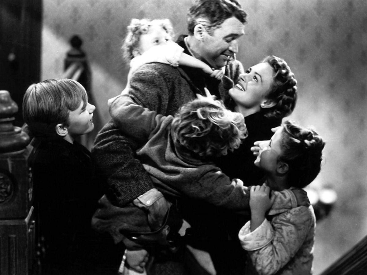 A joyful family embraces on a staircase, with a man and woman smiling at each other while surrounded by four happy children hugging them. The scene is in black and white.