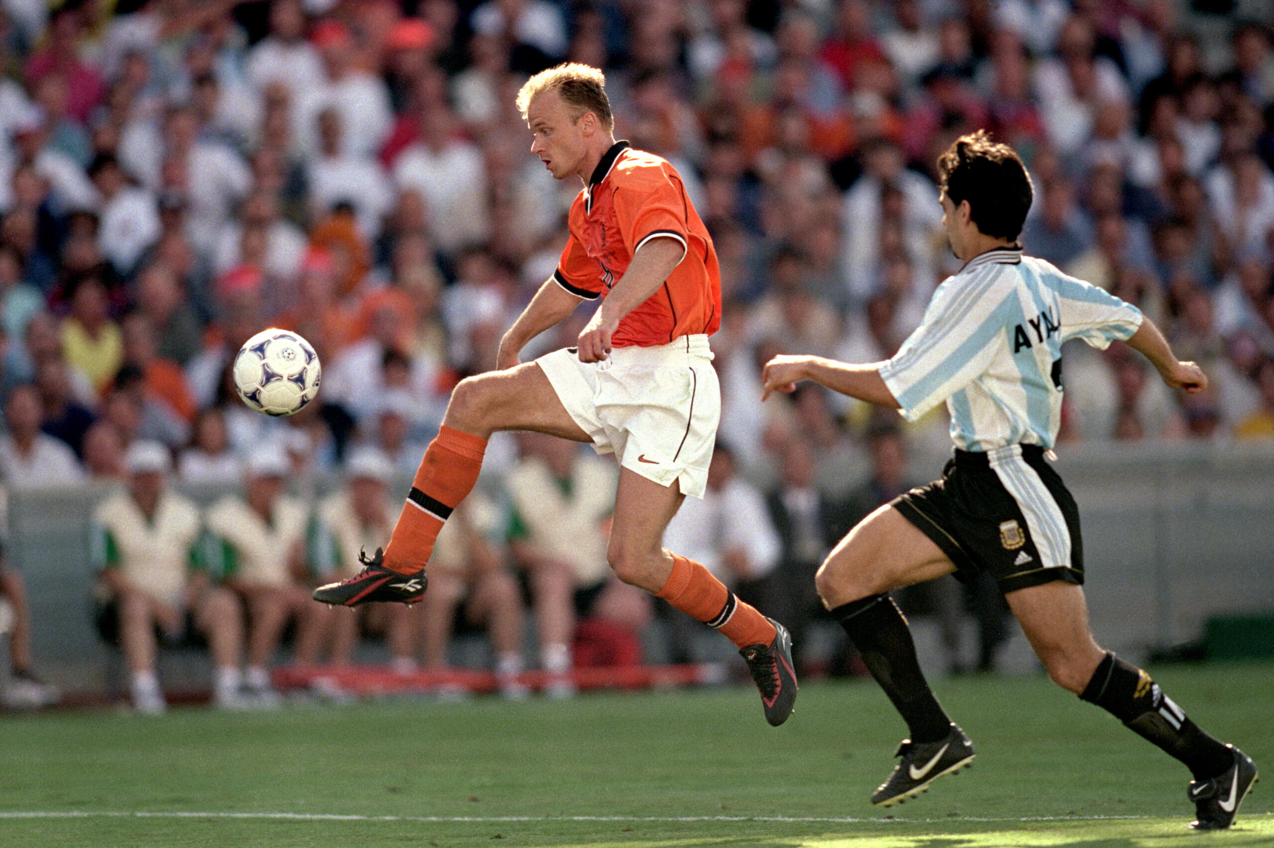 A soccer player in an orange jersey jumps to control the ball mid-air while a player in a light blue and white striped jersey runs behind him on a green field, with spectators in the background.